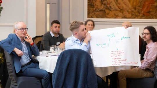 A group of people at a research conference sitting down and listening with one person holding a large piece of paper with data on it