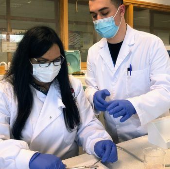 A man and a women are both in a laboratory wearing protective gear, with mask, lab coat and gloves, preparing an experiment.