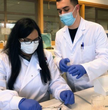 A man and a women are both in a laboratory wearing protective gear, with mask, lab coat and gloves, preparing an experiment.