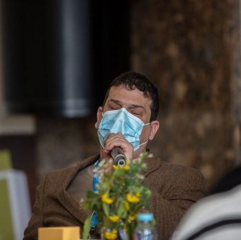 Man speaking at a conference event. He is wearing a mask, while holding a microphone.